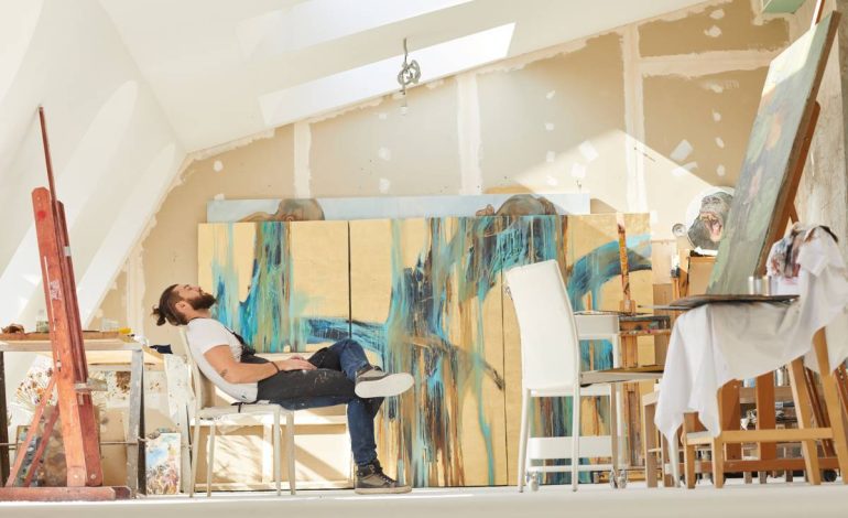Wide angle view at contemporary bearded artist sitting on chair in spacious attic art studio and relaxing after finishing work, copy space